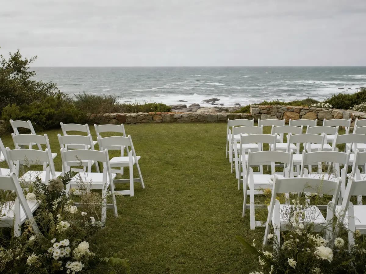 Empty rows of chairs on a coastal lawn in Cape Town with the sea visible beyond a low stone wall