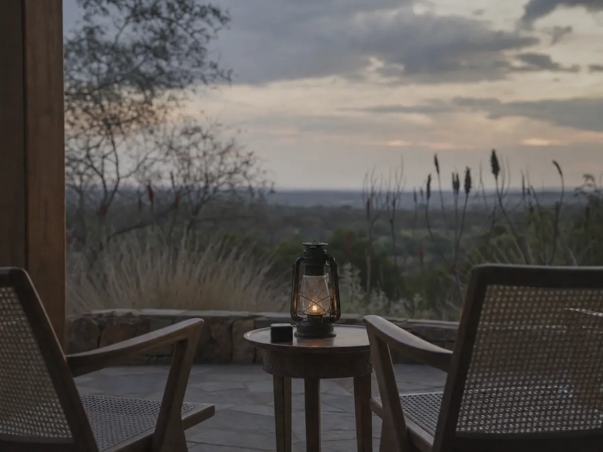 Two chairs on a Highveld stoep at dusk, a lantern between them and a storm clearing to the east
