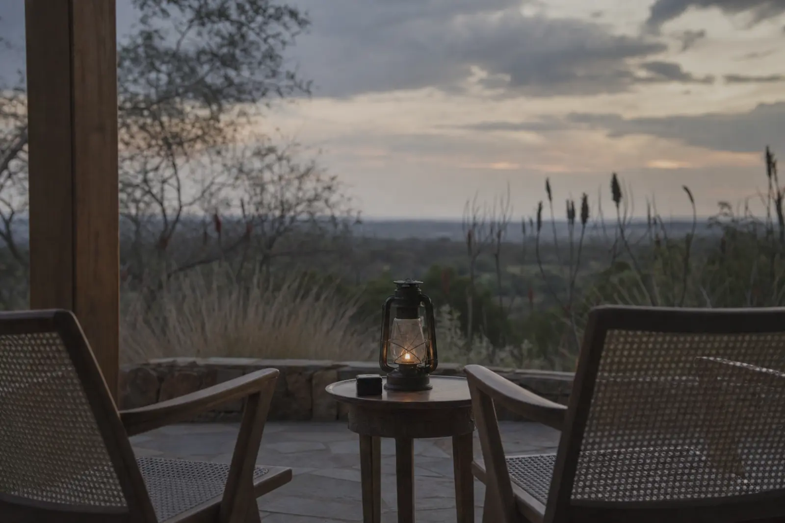 Two chairs on a Highveld stoep at dusk, a lantern between them and a storm clearing to the east