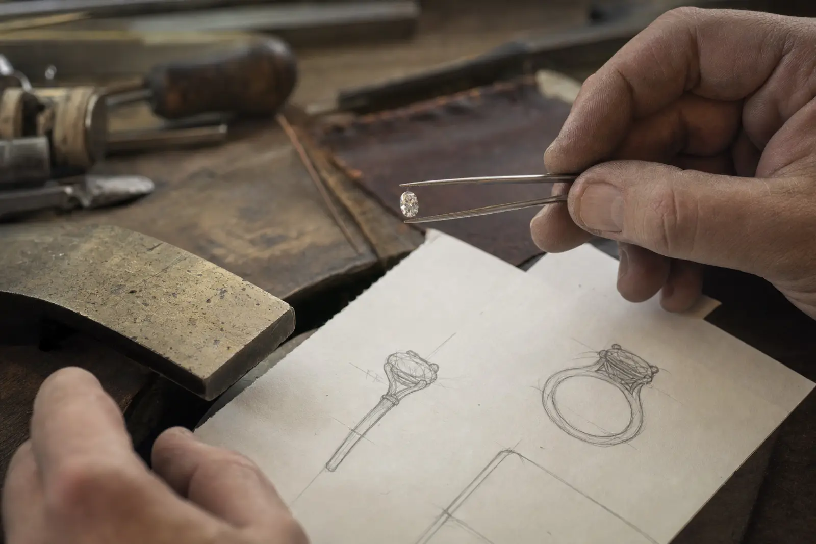 A loose diamond in tweezers above a bench pin, a pencil sketch of its setting beneath it in warm lamp light
