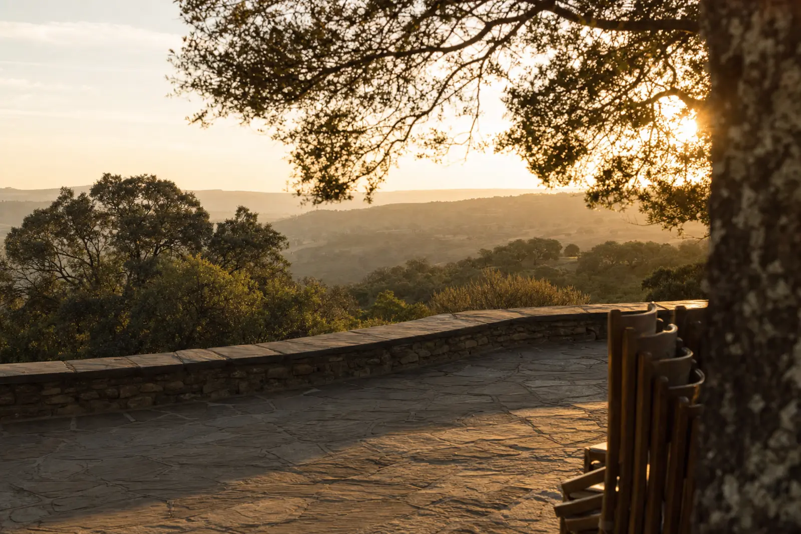 An empty stone terrace above a valley in the late afternoon, chairs stacked to one side and the light going gold