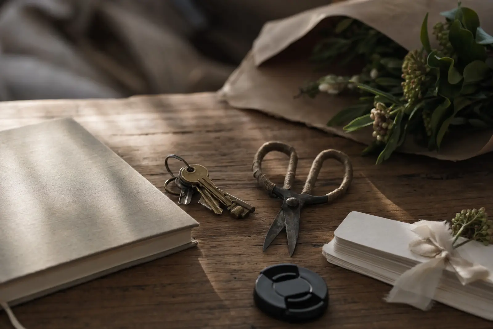 A long wooden table with a planner's notebook, a set of keys, a florist's snips and a photographer's lens cap laid out in a row