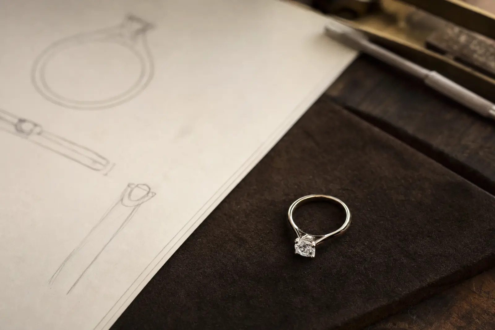 An engagement ring alone on dark suede, drawn with a faint pencil outline of a future band beside it