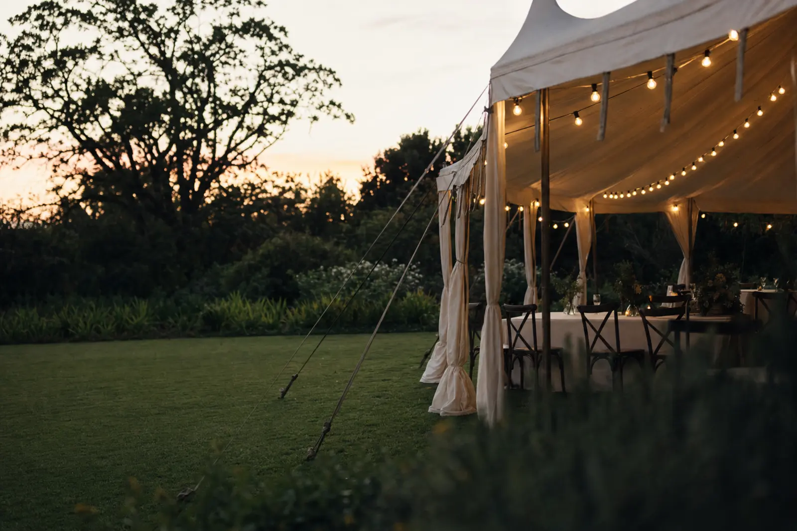 A garden marquee edge with string lighting at dusk, lawn and trees beyond, no people