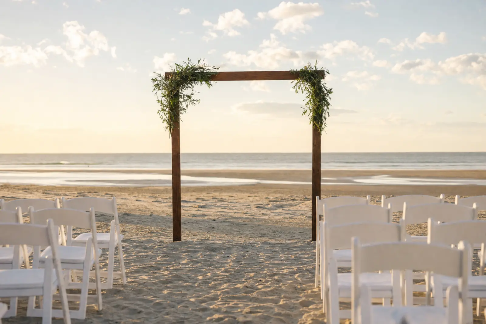 A simple wooden arch on open sand with rows of white chairs facing the sea at low tide