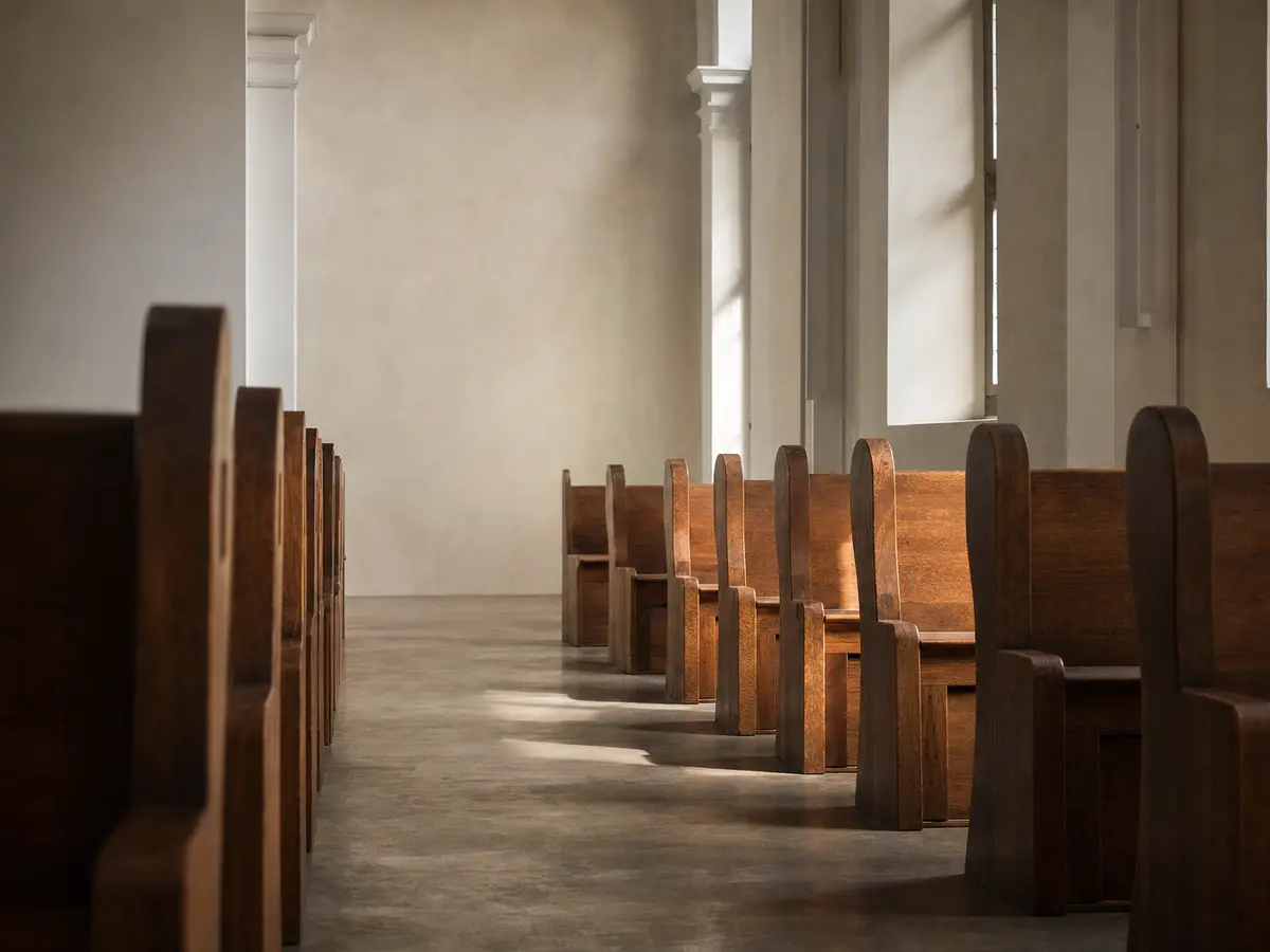 An empty church aisle lined with plain wooden pews, morning light through tall clear windows, no people