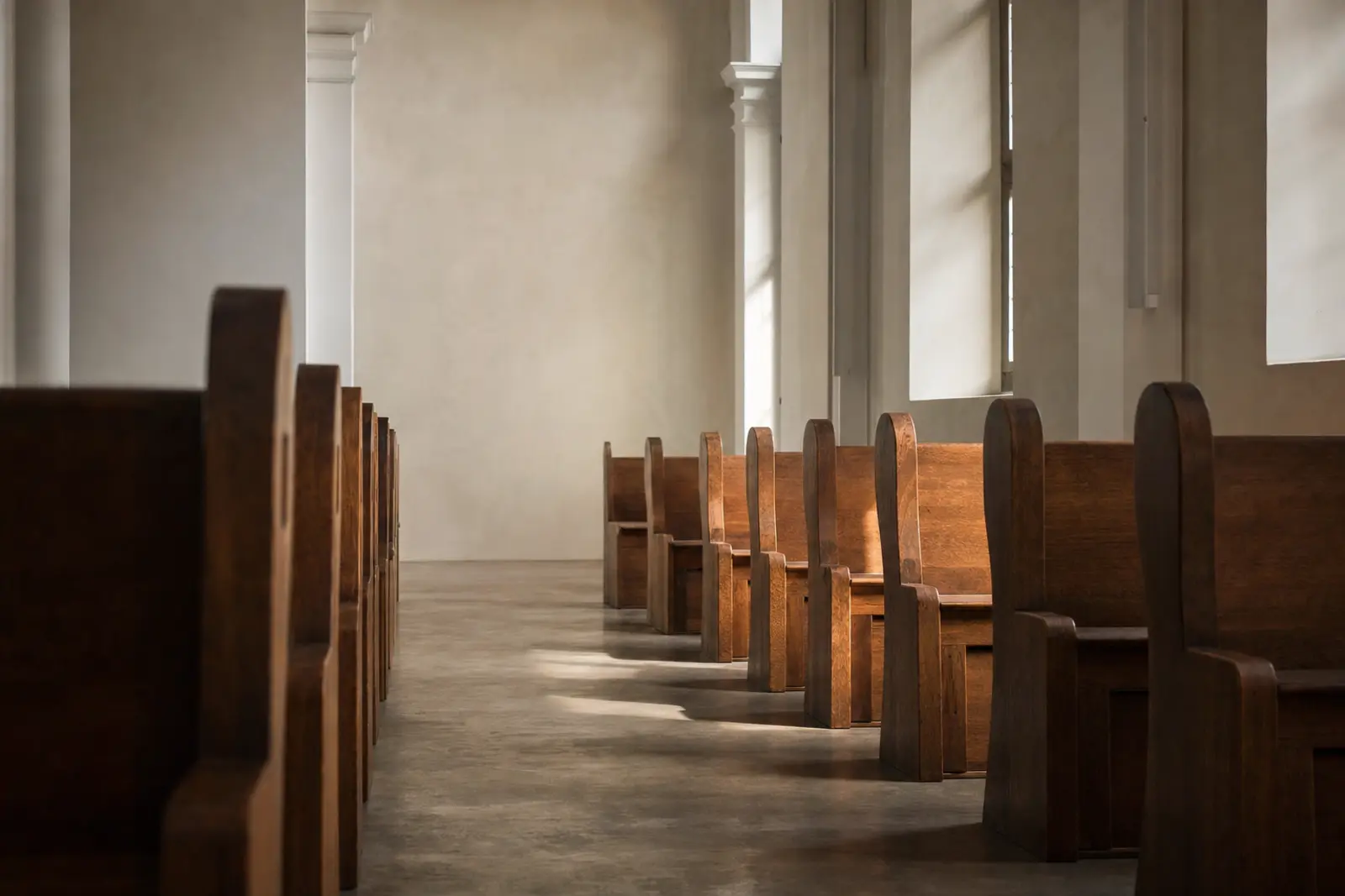 An empty church aisle lined with plain wooden pews, morning light through tall clear windows, no people