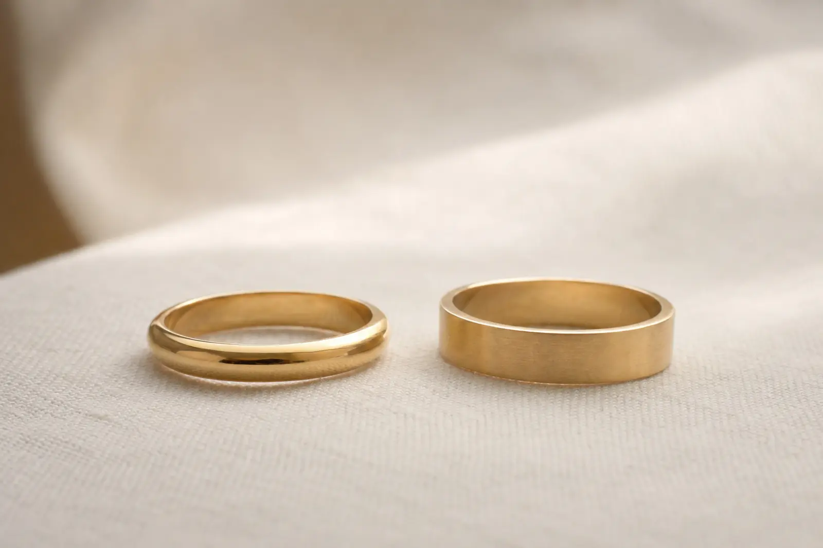 A rounded court-profile wedding band and a flat D-shape band resting side by side on pale linen