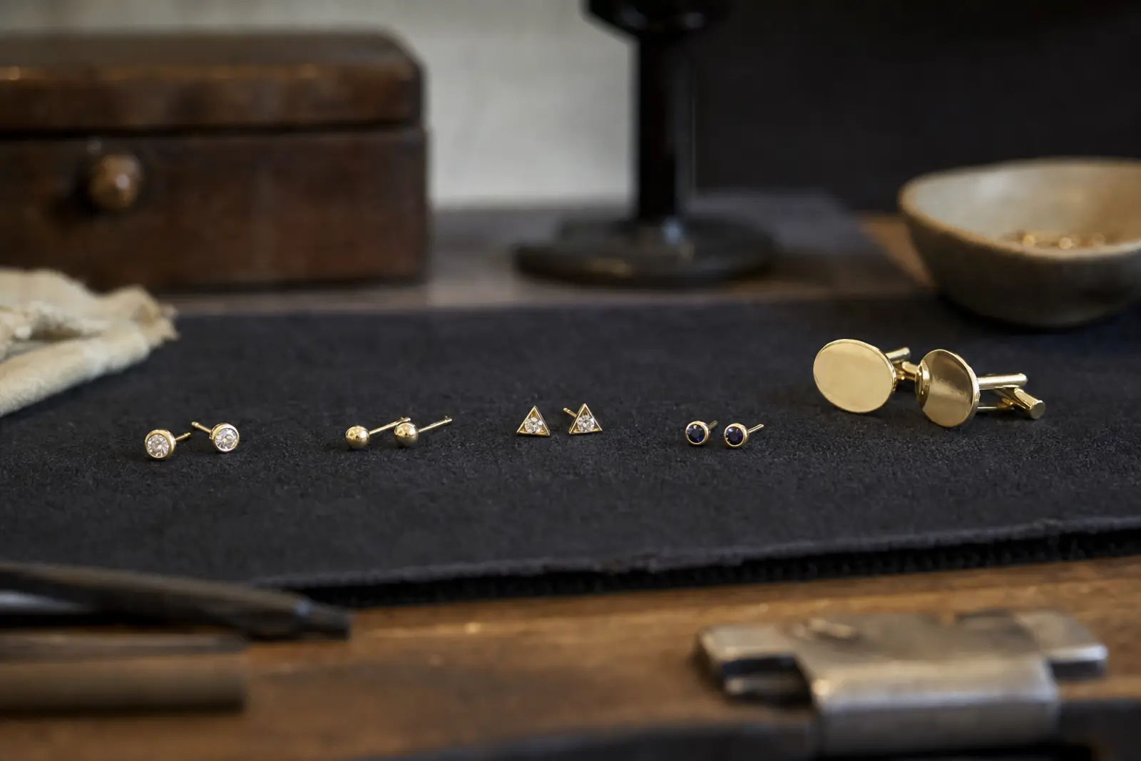 A row of small gold stud earrings and plain cufflinks laid out on a dark cloth